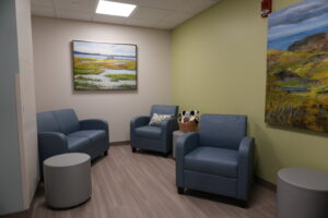 Waiting room with blue upholstered chairs around a small round table, pale walls, and wall art including landscape paintings.