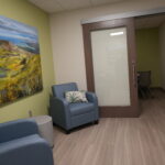 Cozy waiting area with blue upholstered chairs, a large landscape painting on a green wall, and a frosted glass door with a wooden frame.