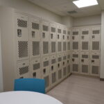 Row of beige metal lockers with ventilation panels along a wall, lime-green wall on the right, and a blue chair in the foreground.