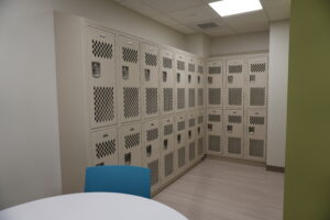 Row of beige metal lockers with ventilation panels along a wall, lime-green wall on the right, and a blue chair in the foreground.