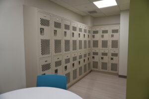 Row of beige metal lockers lining a school hallway, with a blue chair in the foreground and a green wall to the right.