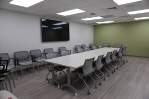 Conference room with a wall-mounted large TV, rows of gray chairs around a long white table, bright ceiling lights.