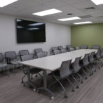 Conference room with a long white table, many gray rolling chairs, and a wall-mounted flat-screen TV on a pale wall with a green accent wall nearby.