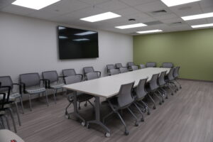 Conference room with a long white table, many gray rolling chairs, and a wall-mounted flat-screen TV on a pale wall with a green accent wall nearby.