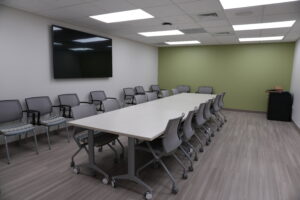 Conference room with a long white table, gray rolling chairs, a large wall monitor, and a green accent wall.