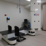 Medical imaging room with a large robotic X-ray table and overhead gantry; sign reads 'Kosair for Kids' on the machine column.