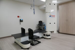 Medical imaging room with a large white scanner, a patient table in front, and a Kosair for Kids sign on a pillar to the right; cables and equipment nearby.