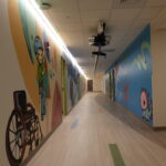 Long hallway with colorful cartoon murals along the walls and a ceiling-mounted projector apparatus overhead.