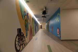 Long hallway with colorful cartoon murals along the walls and a ceiling-mounted projector apparatus overhead.