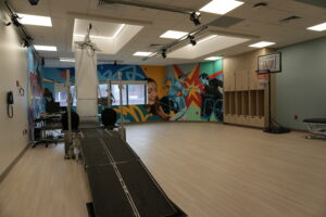 Bright exercise room with a long black treadmill-like track, colorful mural of athletes on the back wall, and a basketball hoop in view.