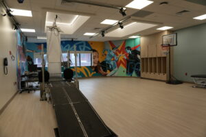 Rehabilitation gym with a long gait-training treadmill, overhead suspension rig, and colorful mural walls on each side.