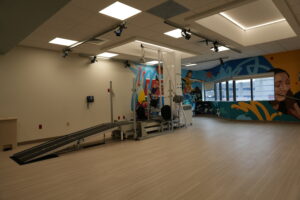 Rehabilitation gym with a harness system spanning overhead, parallel bars, and colorful mural walls along the window wall showing athletes and abstract art.