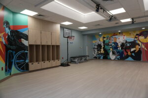 Colorful muraled recreation room with wooden lockers lining the left wall, a small basketball hoop in the center, and bright ceiling lights.