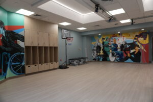 Rehabilitation room with wooden lockers on the left and a colorful mural of people, including a wheelchair user, on the walls; a basketball hoop stands in the center. overhead lights illuminate the room.