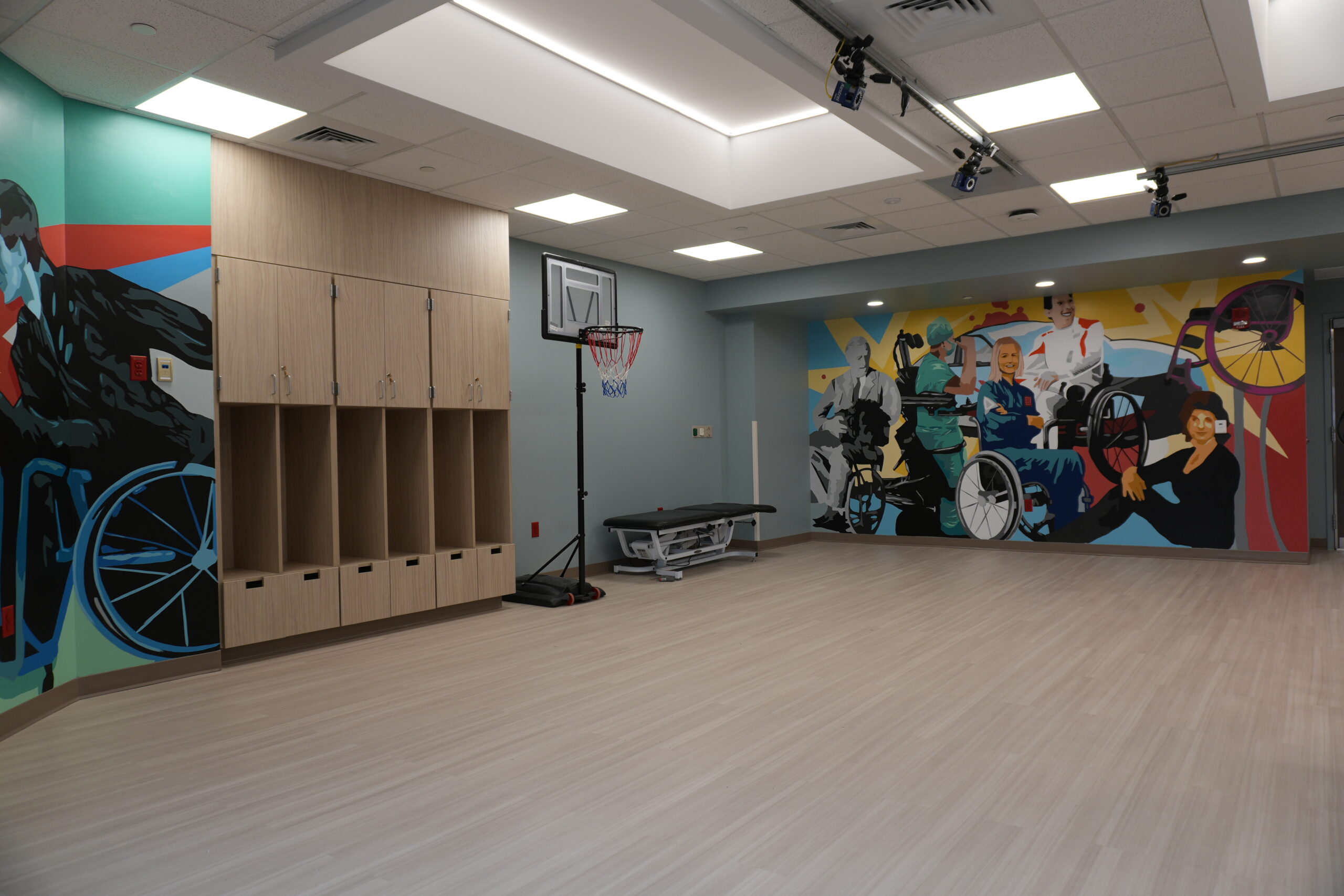 Rehabilitation room with wooden lockers on the left and a colorful mural of people, including a wheelchair user, on the walls; a basketball hoop stands in the center. overhead lights illuminate the room.
