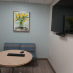 Round conference table with a keyboard and mouse, blue patterned seating, and a framed yellow tulips painting on a pale blue wall; a wall-mounted TV to the right.
