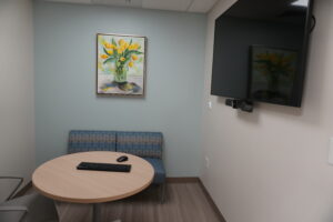 Round conference table with a keyboard and mouse, blue patterned seating, and a framed yellow tulips painting on a pale blue wall; a wall-mounted TV to the right.