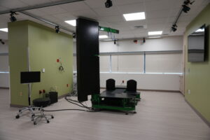 Laboratory room with a large black and green motion/vibration testing machine at center, cables and monitors nearby, green walls, and ceiling lights.