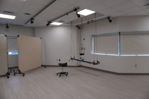 Room with overhead rail system and multiple hanging straps for a harness, plus a black swivel chair in the center foreground and light wood flooring to the left and right.