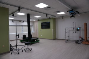 Industrial-looking lab or studio space with ceiling-mounted equipment and rigging, a wall-mounted TV on a green accent wall, and a large metal frame on the right side.