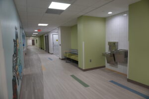 Long hallway with lime-green walls, a drinking fountain area on the right, seating along a wall, and a mural on the left; ceiling lights and an exit sign visible in the distance.