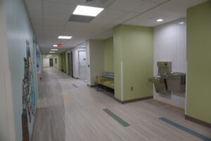 Wide hospital hallway with green walls, a mural on the left, and a water fountain near the right foreground.