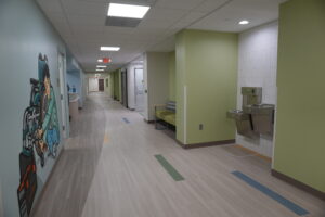 Long hospital hallway with a mural on the left and seating area on the right, extending toward a door in the distance.