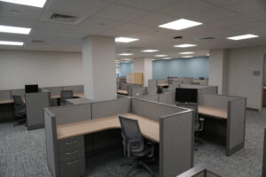 Open-plan office with gray fabric cubicles, wood desks, and ergonomic chairs under a grid ceiling with bright fluorescent lights.