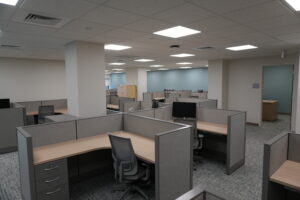 Open-plan office with rows of gray cubicles, beige desks, and swivel chairs under bright ceiling lights; blue wall in the background.