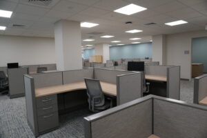Open-plan office with rows of gray cubicles, beige desks, and empty chairs under a drop ceiling.
