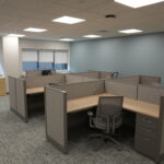 Open-plan office with gray cubicles, beige desks, and gray chairs under a drop ceiling and fluorescent lights; blue-gray accent wall on the right.