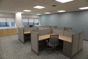 Open-plan office with gray cubicles, beige desks, and gray chairs under a drop ceiling and fluorescent lights; blue-gray accent wall on the right.