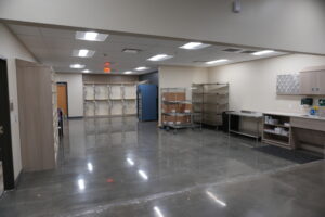 Empty storage room with metal shelving, rolling carts, and boxed supplies along beige walls served by bright overhead lights.