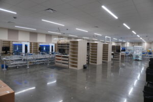 Large open factory floor with conveyor systems, blue-lit control cabinets, and tall wooden shelving units under bright overhead lights