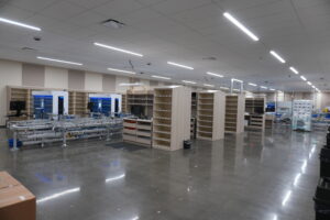 Empty, well-lit industrial workspace with a conveyor assembly line and tall beige shelving units arranged in rows; glossy concrete floor reflects overhead fluorescent lights.