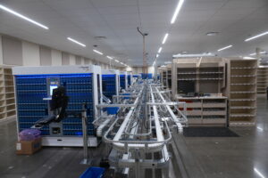 Long row of blue Parata Max 2Plus tablet filling machines with conveyor belts in a clean, well-lit industrial workspace behind empty shelving.