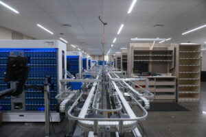 Automated pharmacy belt system with blue Parata machines and empty wooden shelves in a bright warehouse.