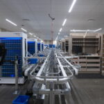 Automated warehouse conveyor system with blue storage racks and empty beige shelving on the right under bright ceiling lights.