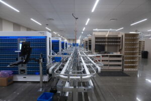 Automated warehouse conveyor system with blue storage racks and empty beige shelving on the right under bright ceiling lights.