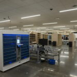 Large warehouse with blue automated storage cabinets, workstations, and overhead fluorescent lights, with staff in the background.