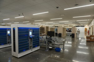 Large warehouse with blue automated storage cabinets, workstations, and overhead fluorescent lights, with staff in the background.