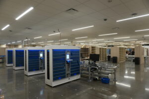 Spacious data-center floor with blue battery racks, conveyor rails, and workstations; a few people in the background near shelving units.