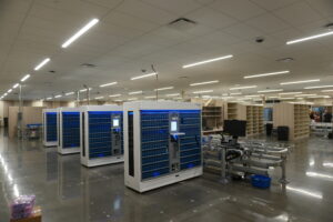 Open data center with white and blue server racks, bright overhead lights, and workstations in the background.