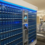 Automated blue-bin storage system with a central touchscreen panel in a lab-like workspace.
