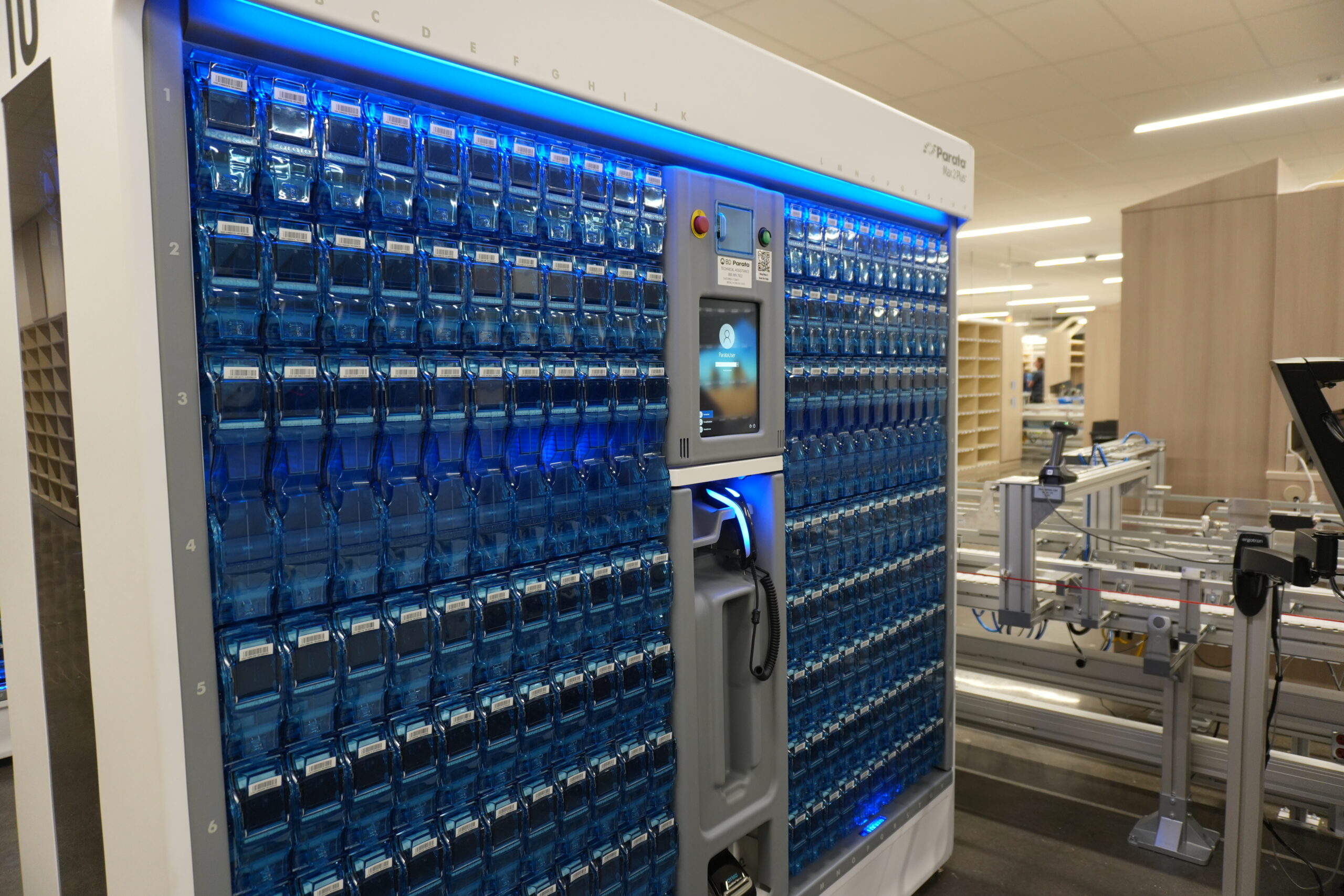 Automated blue-bin storage system with a central touchscreen panel in a lab-like workspace.