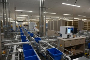 Industrial fulfillment line with conveyor belts and blue plastic bins in a warehouse setting used for automated sorting/packing.