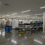 Spacious warehouse interior with tall shelves, carts, and conveyor belts under bright linear lights.
