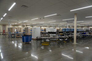 Spacious warehouse interior with tall shelves, carts, and conveyor belts under bright linear lights.