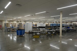 Large warehouse/distribution center with packing stations, shelving, and conveyors under bright overhead lights.