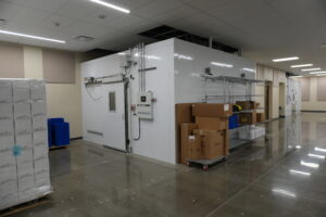 Industrial cold storage room with a large white walk-in freezer, metal shelving, and stacked boxes on carts and pallets nearby.